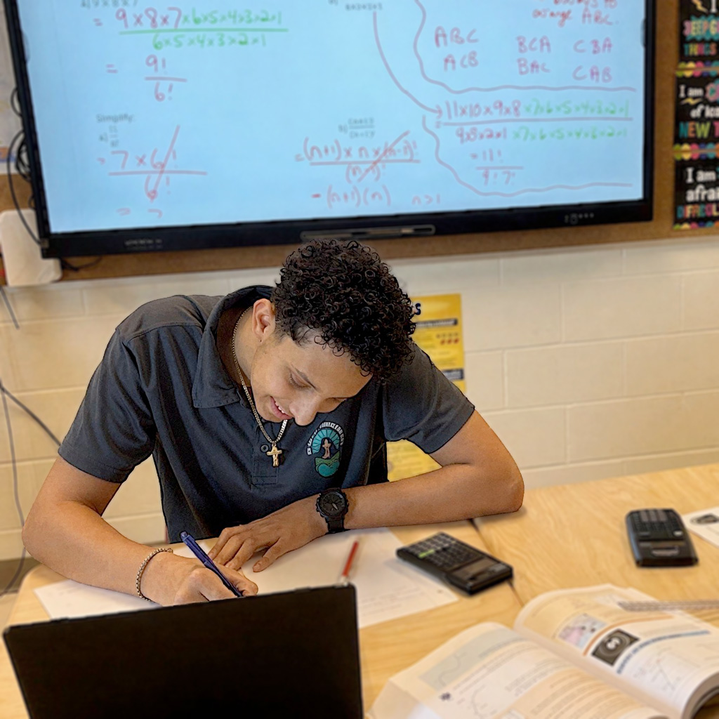 St. Kateri Grade 10 Pre-IB male student working on math equation with whiteboard in background and open laptop and calculator in front of him